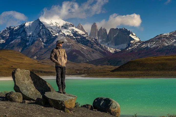 Hombre de pie sobre una roca mirando un lago turquesa y picos irregulares